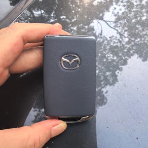 Peut inclure: Un porte-clés de voiture Mazda rectangulaire doté d'une finition texturée gris foncé semblable à du cuir sur la face avant. Le centre du porte-clés affiche un emblème logo Mazda métallique argenté et poli. Les bords ont une finition chromée brillante.
