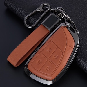 Peut inclure: Un étui de clé en cuir beige avec une lanière en cuir assortie et un mousqueton en métal foncé. L'étui présente une texture lisse avec des icônes en relief pour le verrouillage, le déverrouillage et l'alarme, entouré d'un bord métallique.
