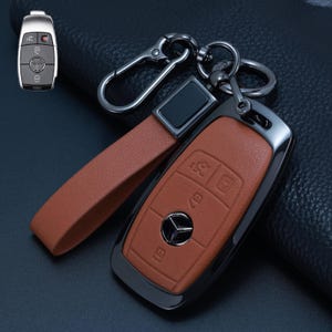 May include: A tan leather key fob cover for a Mercedes-Benz vehicle, featuring a metallic black frame, a matching tan leather wrist strap, and a dark metal carabiner clip. The cover includes embossed button icons and a central circular logo cutout.
