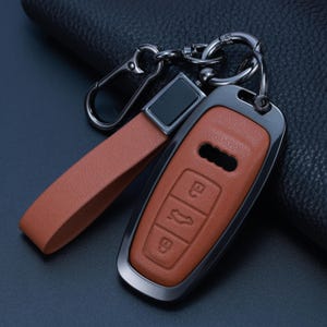 Può includere: Un coprichiave in pelle color cuoio con cornice metallica scura e cinturino da polso abbinato. La custodia presenta icone in rilievo per blocco, sblocco e apertura bagagliaio, oltre a un ritaglio per il logo Audi. È presente un anello portachiavi scuro.
