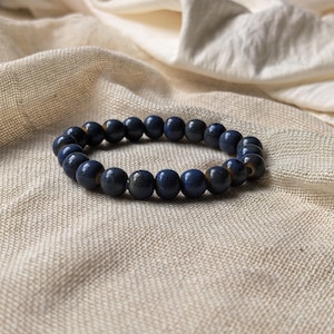 Puede incluir: Una pulsera de cuentas azul marino. La pulsera está hecha de cuentas redondas de color azul oscuro. Las cuentas están ensartadas para formar una forma circular. La pulsera descansa sobre una tela beige.