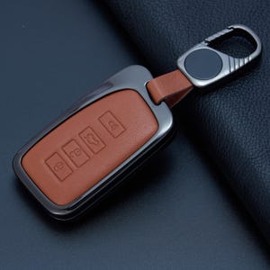 May include: A rectangular car key fob cover featuring a tan leather insert encased in a dark metallic frame. The leather surface displays four embossed icons representing vehicle functions. A matching dark metal carabiner clip is attached by a leather strap.
