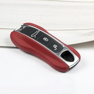 Peut inclure: Une housse de clé de voiture rouge avec une finition texturée lisse et un panneau central noir comportant trois icônes blanches représentant l'ouverture du coffre, un cadenas fermé et un cadenas ouvert, bordée d'un cadre métallique argenté.