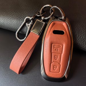 Peut inclure: Un étui de clé de voiture en cuir orange brûlé avec une dragonne en cuir assortie et un porte-clés en métal argenté. L'étui présente des icônes en relief pour le verrouillage, le déverrouillage et le coffre, bordé d'un métal argenté poli.