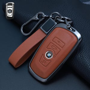 Könnte beinhalten: Eine hellbraune Autoschlüsselhülle aus Leder mit einem metallischen, anthrazitfarbenen Rahmen und einem passenden Lederband. Die Hülle zeigt geprägte Tasten-Symbole und einen kreisförmigen Ausschnitt, der das BMW-Logo freigibt. Ein Karabiner ist befestigt.