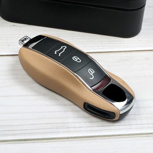 Peut inclure: Une housse de clé de voiture en cuir beige avec un panneau central noir comportant trois icônes blanches représentant un coffre, un cadenas fermé et un cadenas ouvert. Le boîtier présente une bordure argentée, un anneau métallique et une base noire.