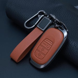 May include: A tan leather car key fob cover with a matching leather strap and a dark metallic carabiner clip. The cover features embossed icons for locking, unlocking, and a hold function, encased in a sleek, dark-toned metal frame.