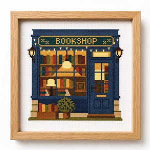 Puede incluir: Un bordado de punto de cruz de la fachada de una librería en un marco de madera clara. El diseño presenta una fachada azul oscuro, un letrero que dice BOOKSHOP, grandes ventanales con libros coloridos, una puerta, dos lámparas y una planta.
