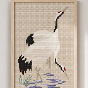 May include: Cross-stitch artwork of two Japanese cranes in a natural setting. The birds are white with black wingtips and red accents. The background is a neutral beige, and the artwork is framed in light wood.