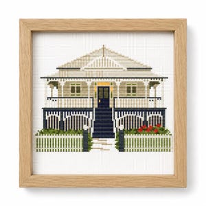 May include: A cross-stitch embroidery of a house with a beige exterior, a dark blue door, and a dark blue foundation. The house features a white picket fence, green bushes with red flowers, and a light-colored roof, all encased in a light wood frame.