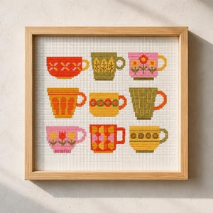 May include: A framed cross-stitch artwork featuring nine colorful teacups. Each cup has a unique design in shades of red, green, yellow, and pink. The artwork is set against a neutral background and framed in a light wood.