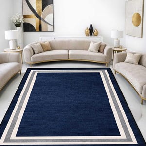 May include: A rectangular navy blue area rug with a gray and white border. The rug is placed in a living room with neutral-toned furniture and artwork. The rug's dimensions are not visible.