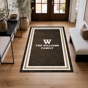 May include: A rectangular brown rug with a white border and a tan stripe. The rug features the letter "W" and the words "THE WILLIAMS FAMILY" in white. The rug is placed on a wooden floor.