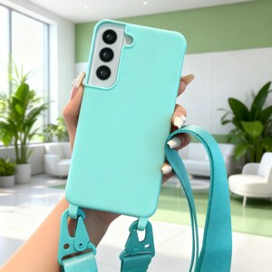 May include: A light teal phone case with a matching strap. The case has a smooth finish and features three camera lenses. The strap is attached with teal-coloured clips. The phone case is held by a person.
