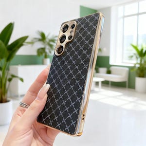 May include: A black phone case with a gold-coloured frame and camera module. The case features a textured, cross-patterned design. The phone case is held in a person's hand.