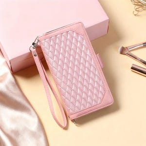 Puede incluir: Un monedero de cuero acolchado de color rosa claro con una correa de muñeca. El monedero tiene un patrón de diamantes y un cierre de cremallera. El fondo incluye una caja rosa, brochas de maquillaje y un clip dorado.