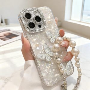 Può includere: Una custodia trasparente per telefono con un design di perle e farfalle. La custodia presenta un cinturino di perle ed è decorata con farfalle scintillanti e piccole perle. L'obiettivo della fotocamera è circondato da una cornice argentata tempestata di gemme.