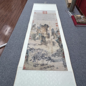 May include: A long Chinese scroll painting depicting a landscape with mountains, trees, and buildings. The artwork uses grey, black, and brown tones, with red seals and calligraphy. The scroll is rolled at the bottom with wooden dowels, and is approximately 150 cm long.