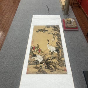 Könnte beinhalten: Traditionelle chinesische Rollbildmalerei mit zwei Kranichen in einer Landschaft aus Bäumen und Blumen. Das Kunstwerk verwendet Gelb-, Braun- und Grüntöne, mit weißen und schwarzen Kranichen. Die Schriftrolle hat einen weißen Rand und eine dunkelrote Holzrolle.
