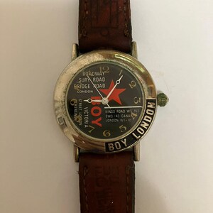 May include: A wristwatch with a dark brown leather strap and a silver-tone metal case. The black watch face features a red star, white text reading ROADWAY SURY ROAD BRIDGE ROAD LONDON, BOY, and KINGS ROAD W5 153 SW3-43 CANAR LONDON W1-10.