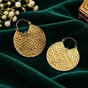 May include: A pair of circular gold hoop earrings featuring a textured surface with an intricate geometric pattern of squares and lines. Each earring has a notched top section and a thin wire closure, showcasing a metallic finish and detailed relief.