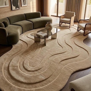 May include: A large, beige, irregularly shaped area rug featuring a raised, wavy, concentric line pattern that creates a layered, topographical effect across its surface. The rug is positioned on a dark wooden floor beneath a glass coffee table.