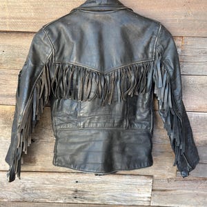 Puede incluir: Una chaqueta de cuero negro con flecos largos en la espalda y a lo largo de las mangas. La prenda presenta un canesú trasero en punta, un cuello estructurado y mangas largas con puños de cremallera, mostrando una textura desgastada.