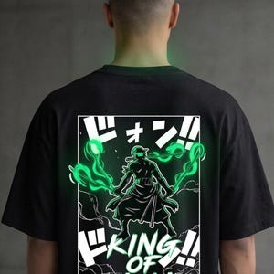 Puede incluir: Camiseta negra con un gráfico blanco y verde de un personaje con espadas y el texto "KING OF HELL". El diseño tiene un efecto de brillo verde neón. Una prenda casual.
