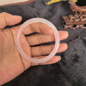 Puede incluir: Un brazalete de jade rosa pálido. El brazalete circular se sostiene en una mano sobre un fondo gris oscuro. El brazalete tiene un aspecto pulido y translúcido, reflejando la luz. Una pieza de joyería tradicional.
