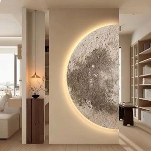 May include: A large, textured, half-moon wall art piece with a warm, glowing halo. The moon's surface is a mix of gray and beige, with craters and a rough texture. The art is set against a neutral wall, creating a focal point in a modern interior.