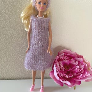 Könnte beinhalten: Eine Puppe trägt ein flauschiges lavendelfarbenes Kleid, einen passenden Hut und rosa Schuhe. Die Puppe hat blondes Haar und eine weiße Halskette. Eine große rosa Blume befindet sich neben der Puppe.