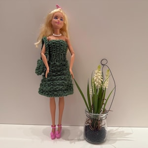 Könnte beinhalten: Eine Puppe in einem handgefertigten grünen Strickkleid und einer passenden Handtasche, kombiniert mit rosa Absätzen. Die Puppe hat blondes Haar und eine Perlenkette. Ein kleines Glas mit weißen Blumen und Grünzeug ist ebenfalls im Bild.