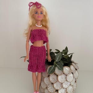 May include: A blonde doll with blue eyes wearing a magenta knitted two-piece outfit consisting of a strapless tube top and a matching skirt. The doll has a pink hair tie, a white beaded necklace, and pink sandals. The doll is standing upright.
