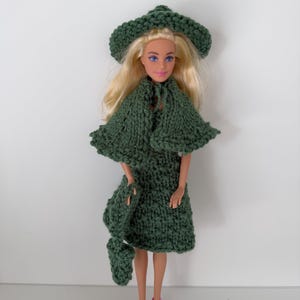 Könnte beinhalten: Eine Puppe in einem handgefertigten, grünen Strick-Outfit, einschließlich Hut, Umhang, Kleid und Schal. Die Puppe hat blondes Haar und trägt rosa Schuhe. Das Outfit hat eine einfarbige, dunkelgrüne Farbe.