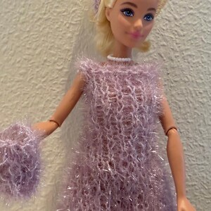Könnte beinhalten: Eine Puppe in einem hellvioletten, flauschigen Kleid und passenden Handgelenkmanschetten. Die Puppe hat blondes Haar und trägt eine weiße Halskette und ein passendes Stirnband. Das Kleid ist ärmellos.