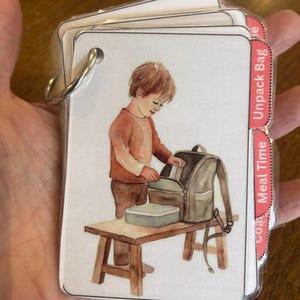 Puede incluir: Un conjunto de tarjetas ilustradas unidas por un anillo metálico. Cada tarjeta presenta una ilustración en acuarela de un niño. Las tarjetas tienen pestañas rosas con texto, incluyendo "Unpack Bag" y "Meal Time".