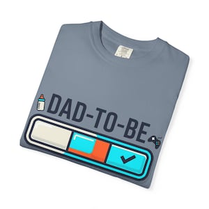 Può includere: Una maglietta blu ardesia con la scritta "DAD-TO-BE" e una grafica di un biberon, una barra di avanzamento e un controller di videogiochi. La barra di avanzamento ha sezioni bianche, arancioni e blu, con un segno di spunta.