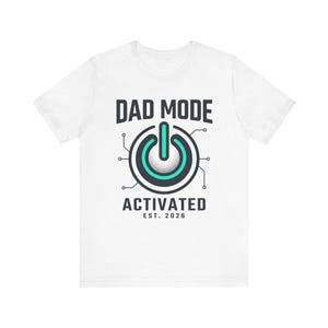 May include: White t-shirt with the text "DAD MODE ACTIVATED EST. 2026". The design features a power button graphic with a teal center and black outline, surrounded by circuit-like lines.