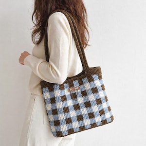 May include: A brown, blue, and white checkered crochet tote bag with brown handles. The bag has a rectangular shape and a small leather tag. The bag is carried over the shoulder.