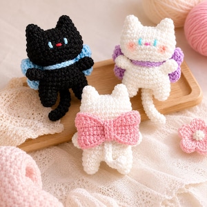 May include: Three handmade crochet cat figurines. One black cat with blue accents, a white cat with purple wings, and a white cat with a pink bow. A pink crochet flower is also visible. The cats are on a wooden tray.