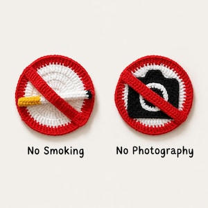 Puede incluir: Dos letreros circulares de ganchillo con bordes rojos y una franja diagonal roja. El letrero izquierdo presenta un centro blanco con un cigarrillo y el texto No Smoking. El derecho muestra una cámara negra en el centro y el texto No Photography.