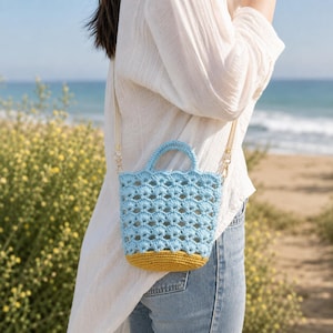 Pode incluir: Uma bolsa de mão em crochê azul-claro com um padrão de vieiras e uma base sólida em amarelo-mostarda. A bolsa possui duas alças pequenas superiores e uma alça de ombro removível na cor creme com clipes de metal dourado nas laterais.
