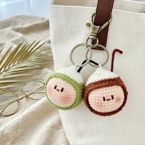May include: Two handmade crochet acorn keychains. One keychain has a green cap, the other has a brown cap. Both have white tops, pink cheeks, and small black eyes. The keychains are attached to a silver keyring and a white tote bag.