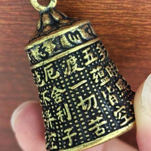 May include: A small, antique-style bell with a loop at the top. The bell is dark bronze with gold accents and is covered in intricate Chinese characters. The text "can produce sound!" is at the bottom.