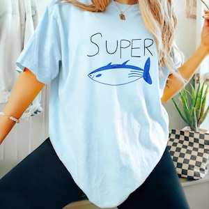 Op de afbeelding: Lichtblauw T-shirt met het woord "SUPER" in zwarte letters boven een blauwe visillustratie. De vis heeft een gestroomlijnd lichaam met vinnen en een staart. Het shirt is een casual, comfortabele stijl.