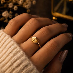 Op de afbeelding: Een goudkleurige ring met de letter "S" gegraveerd op een decoratieve, schildvormige zetting. De ring zit om een vinger, tegen een donkere achtergrond. Een mouw van een neutrale kleur trui is ook zichtbaar.