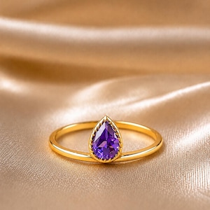 Può includere: Un anello in oro con una singola pietra preziosa viola a forma di goccia incastonata in una lunetta dentellata. La fascia è sottile e lucida, con un profilo liscio e arrotondato che completa la pietra viola sfaccettata al centro.