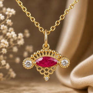 May include: A gold necklace featuring a pendant with a central marquise-cut red gemstone, flanked by two round clear stones. The pendant has a decorative filigree metalwork design with small gold beads along the top edge and a tiny clear stone at the base.