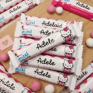 Può includere: Barrette di cioccolato Kinder Maxi Hello Kitty bianche e rosa con il nome "Adele" e "3 anni" stampato sopra. Le barrette sono disposte su un tagliere di legno, con caramelle rosa e bianche.