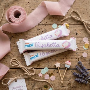 Può includere: Un tavolo di legno mostra barrette di cioccolato Kinder Maxi con il nome "Elisabetta" e un design a unicorno. Nastro rosa, fiocchi di spago, coriandoli e stuzzicadenti decorativi con un cupcake e una stella creano un'atmosfera di compleanno festosa.